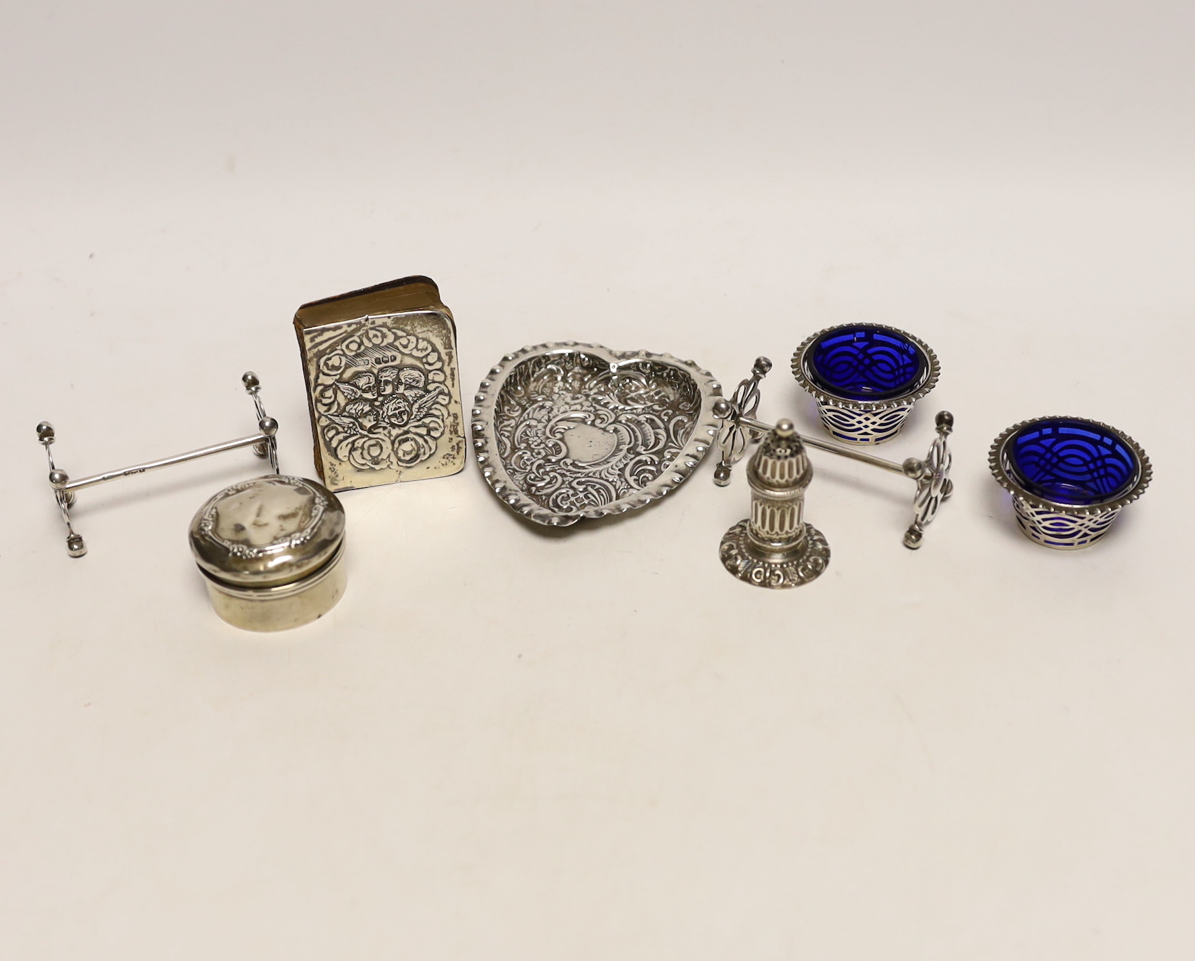 An Edwardian repousse silver mounted small bible, a silver circular pill box, a pair of silver salts, silver pepperette, pair of silver knife rests and a heart shaped dish.